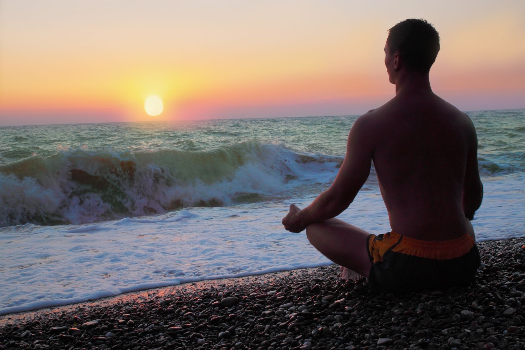  A person sits on the beach in a meditative pose as the sun sets over the ocean.